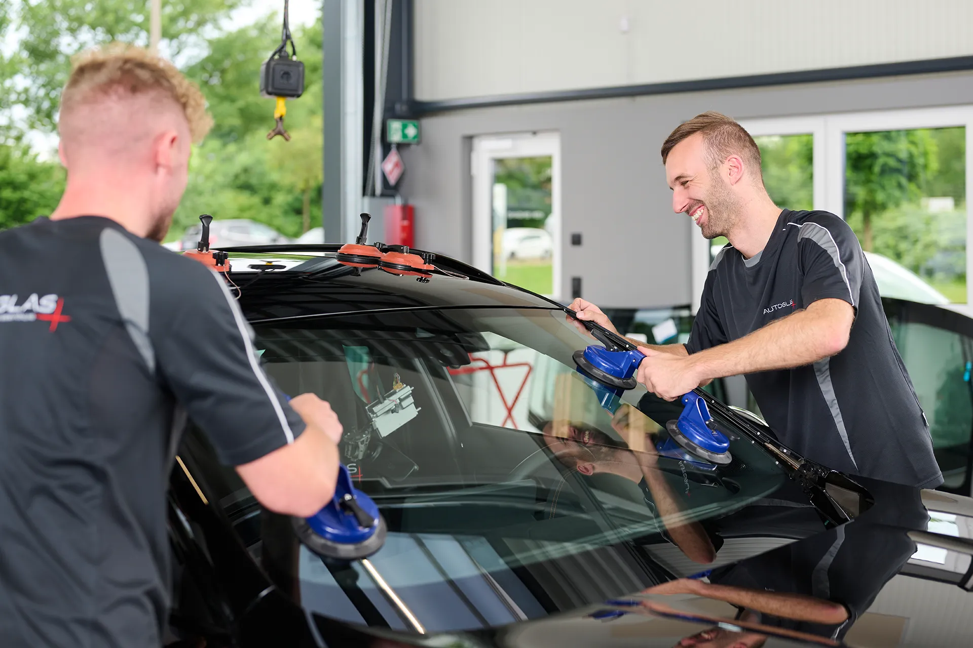Zwei Männer montieren in einer Autowerkstatt mithilfe von Saugnäpfen eine Windschutzscheibe an einem schwarzen Auto.