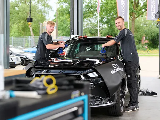 Zwei Männer montieren in einer Autowerkstatt mithilfe von Sauggreifern eine Windschutzscheibe an einem schwarzen Auto.