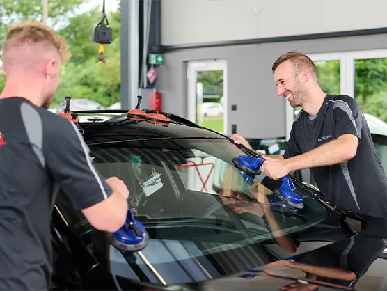 Zwei Männer montieren in einer Autowerkstatt mithilfe von Sauggreifern eine Windschutzscheibe an einem schwarzen Auto.